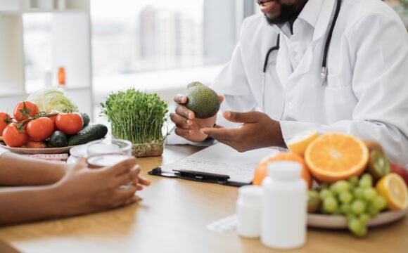 Close up view of bearded man holding green fruit while chatting with multiethnic person in office interior. Male nutritionist describing benefits of slimming foods during appointment in modern clinic.